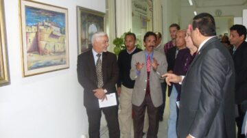 مجموعة من الرجال والنساء يتجمعون في معرض فني، حيث يتم عرض لوحات ملونة على الجدران. يتحدث أحد الرجال بوضوح، بينما يستمع الآخرون بتركيز. يظهر في الخلفية بعض الأعمال الفنية والمناظر المعمارية.