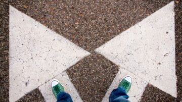 أقدام ترتدي أحذية رياضية خضراء تقف في منتصف علامة مرور بيضاء على شكل سهمين متقابلين على سطح أسفلتي.