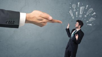 رجل في بدلة رسمية يبدو قلقًا ومرتبكًا بينما تشير إليه يد كبيرة بأصابع ممدودة. الخلفية ذات لون رمادي فاتح مع رسومات توضيحية توحي بالتحذير. تعكس الصورة مشاعر الضغط والتوتر في بيئة العمل.