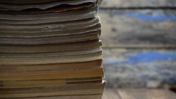 صورة لرف من المجلات القديمة المكدسة، تظهر الصفحات الممزقة والألوان الباهتة، مع خلفية خشبية تعكس جوًا من العراقة والحنين.