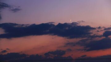 غروب شمس جميل يظهر سماء ملونة بألوان دافئة، مع وجود هلال رقيق في الزاوية العليا. السحب تتناثر في الأفق، مكونة مشهداً طبيعياً ساحراً.