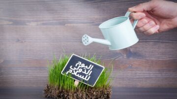 يد تظهر وهي تسقي نباتات خضراء في وعاء مع لافتة مكتوب عليها "اليوم الدولي للسعادة" على خلفية خشبية.