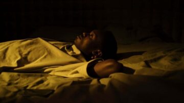 رجل مستلقٍ على سرير في غرفة مضاءة بشكل خافت، يبدو عميق التفكير أو التأمل، بينما يضع ذراعيه خلف رأسه.