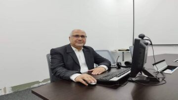 رجل مسن يرتدي بدلة رسمية يجلس على مكتب في مكتب حديث، أمامه جهاز كمبيوتر. يظهر في الخلفية جدار أبيض وخلفية بسيطة.