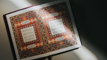 صفحتان مفتوحتان من القرآن الكريم، تظهران النقوش والزخارف الملونة بشكل جميل، مع نص عربي واضح. الإضاءة تسلط الضوء على الكتاب، مما يبرز تفاصيل التصميم والفن الإسلامي.