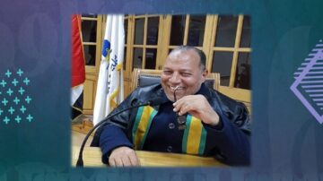 رجل مبتسم يرتدي رداء قضائي، يجلس أمام ميكروفون مع نظارات في يده. تظهر في الخلفية أعلام وجدران خشبية، مما يدل على بيئة رسمية.