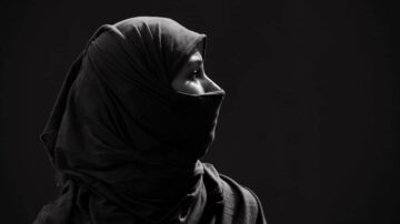 امرأة ترتدي حجابًا أسود، تظهر في صورة بالأبيض والأسود. تركز الصورة على جانب وجهها، مما يبرز ملامحها في إضاءة خافتة. خلفية الصورة داكنة، مما يضيف عمقًا للتركيز على تعبيرها.