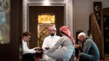 رجلان يجلسان معاً في مسجد، يقرآن القرآن مع معلم يرتدي ثوباً تقليدياً وعقال. الأجواء هادئة ومليئة بالروحانية، مع تفاصيل معمارية إسلامية في الخلفية.