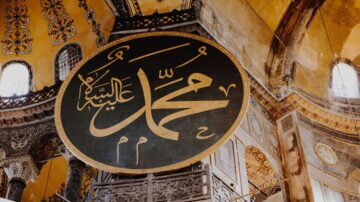 خط كبير باللون الأسود مكتوب عليه "محمد" بخط عربي جميل، يتوسط قبة آيا صوفيا، محاط بزخارف ذهبية وتفاصيل معمارية تاريخية.