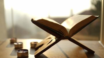 كتاب مفتوح على حامل خشبي، مع شموع صغيرة تحيط به، والإضاءة الطبيعية تبرز تفاصيل الصفحات.