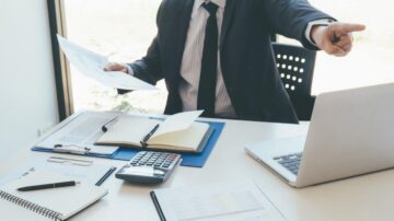 رجل أعمال يرتدي بدلة رسمية يجلس على مكتب، ممسكًا بأوراق في يد، ويشير بإصبع إلى الشاشة أمامه. على المكتب هناك دفتر ملاحظات، قلم، آلة حاسبة، وأوراق مرتبة. الضوء الطبيعي يدخل من نافذة خلفه، مما يخلق أجواء مهنية.