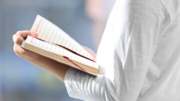 امرأة تحمل كتابًا مفتوحًا، تظهر يدها فقط، بينما الكتاب يحتوي على نص مكتوب. الخلفية غير واضحة، مما يبرز الكتاب.