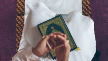 شخص يرتدي الثوب العربي، يمسك بمسبحة في يده، بينما يتواجد القرآن الكريم أمامه على سجادة صلاة.