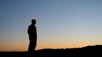 رجل يقف silhouetted ضد سماء غروب الشمس، مع تدرجات اللون البرتقالي والأزرق. المنظر يعكس شعورًا بالهدوء والتأمل في الطبيعة.