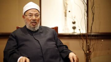 رجل مسن يرتدي عمامة وعباءة في جلسة حوارية، يظهر في خلفية داخلية حديثة مع عناصر زخرفية. تعبيره جدي، مما يعكس موضوعًا جادًا.