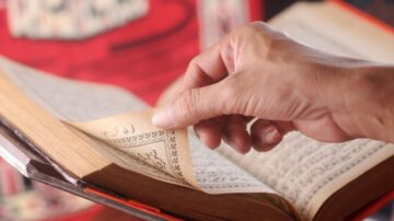 يد يظهر وهو يقلب صفحات كتاب قديم، مع التركيز على النص المكتوب بخط عربي. الخلفية تحتوي على تفاصيل زاهية تعكس التراث الثقافي.