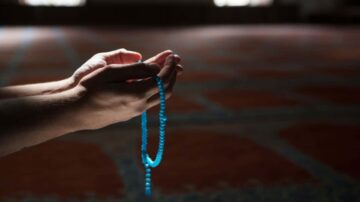 يدان مفتوحتان تحملان سبحة زرقاء في مكان هادئ. الأرضية مزخرفة بنمط تقليدي، مما يضيف جوًا من السكينة والروحانية.