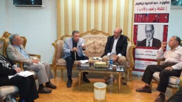 اجتماع جماعي في غرفة جلوس أنيقة، حيث يجلس خمسة أشخاص حول طاولة. يتحدث رجل في المنتصف بجدية، بينما يستمع الآخرون بانتباه. خلفهم يظهر لافتة تتضمن صورة واسم شخصية بارزة، بالإضافة إلى نصوص باللونين الأحمر والأسود. الأثاث فخم ويعكس طابعاً عربياً تقليدياً.