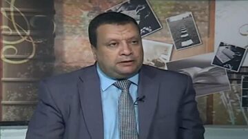 رجل يرتدي بدلة رسمية يجلس أمام خلفية تحتوي على صور فنية وكتب. يبدو في منتصف حديث أو مقابلة تلفزيونية، مع تعبير جاد.