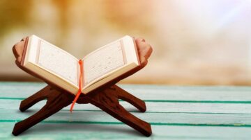 كتاب موضوع على حامل خشبي، يظهر في خلفية ضبابية، مع ضوء طبيعي يتسلل من الجانبين. التصميم يعكس جمال الفن الإسلامي ويبرز تفاصيل الصفحات.