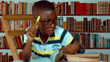 صبي يرتدي نظارات ويمسك بقلم أصفر، يجلس على كرسي ويقرأ كتابًا في مكتبة تحتوي على رفوف مليئة بالكتب.