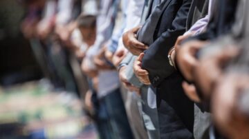 صف من الرجال يؤدون الصلاة في صفوف منتظمة، مع التركيز على أذرعهم المتشابكة. يظهر في الخلفية أشخاص آخرون وهم يقيمون شعائر دينية.