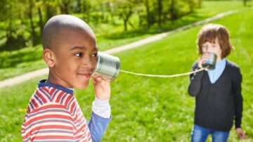 طفلان يلعبان في حديقة، أحدهما يتحدث في علبة معدنية متصلة بخيط، بينما الآخر يستمع. يظهر المشهد تفاعلًا ممتعًا بين الأطفال في بيئة طبيعية.