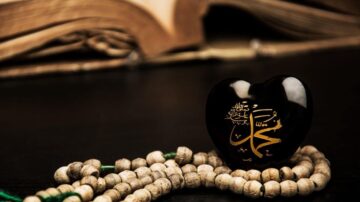 قلب أسود مزين بالخط العربي يحمل اسم محمد، موضوع بجانب سبحة خشبية، مع كتاب قديم في الخلفية. تعبير عن الإيمان والثقافة الإسلامية.