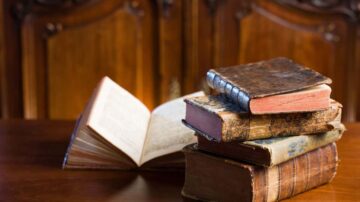 كتب قديمة موضوعة على طاولة خشبية، مع كتاب مفتوح في الخلفية. تظهر تفاصيل جلد الكتب والتجاعيد على صفحاتها، مما يبرز قيمتها التاريخية.