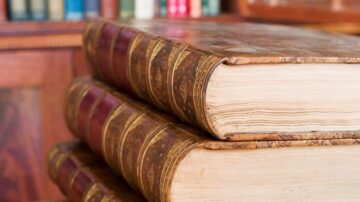 كتب قديمة ذات غلاف جلدي بني، موضوعة فوق بعضها في مكتبة تقليدية، مع رفوف مليئة بالكتب الملونة في الخلفية.