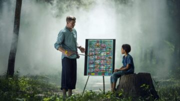 معلم يشرح درسًا لطفل يجلس على جذع شجرة، في غابة ضبابية. خلفهم لوحة تعليمية تحتوي على صور ملونة.