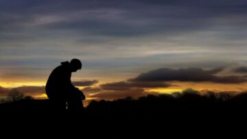 شخص جالس في ظلام مع غروب الشمس في الخلفية، يظهر silhouette للشخصية أمام سماء ملونة بالبرتقالي والأزرق. تعكس الصورة مشاعر التأمل والهدوء.