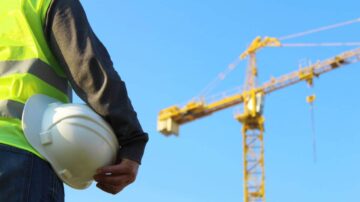 رجل يرتدي سترة عمل وقبعة سلامة، يقف في موقع بناء أمام رافعة عملاقة تحت سماء زرقاء.