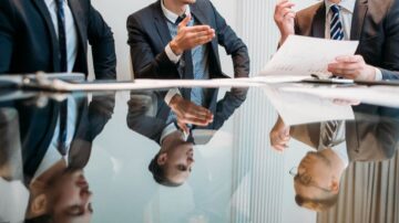 اجتماع عمل يضم ثلاثة رجال يرتدون بدلات رسمية، يجلسون حول طاولة زجاجية. يظهر في الصورة انعكاسهم على السطح الزجاجي، حيث يتحدث أحدهم بيد مرفوعة بينما يراجع الآخرون مستندات. تعكس البيئة المهنية أجواء النقاش والتفاعل.