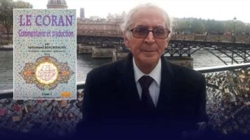 رجل مسن يرتدي نظارات يقف أمام جسر في خلفية نهر مع كتاب "القرآن: تفسير وترجمة" بجانبه. الكتاب يحمل عنوانه باللغة الفرنسية، مع تفاصيل زخرفية.