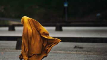 شخص يرتدي عباءة صفراء يخفي جسده، يتجول في منطقة خارجية ذات أرضية حجرية. تظهر الخلفية بلون داكن مع بعض الإضاءة. تعبر الصورة عن الغموض والحركة.