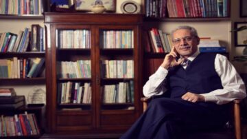 رجل مسن يجلس في كرسي مريح، مرتديًا بدلة رسمية ونظارات. خلفه مكتبة تحتوي على العديد من الكتب. الإضاءة دافئة وتضفي جوًا هادئًا.