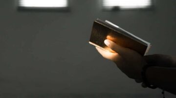 يد شخص يحمل كتابًا في غرفة مضاءة بخافتة، مع تسليط الضوء على الكتاب من جانب اليد.