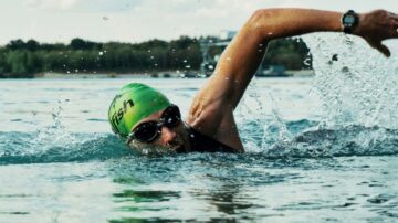 سباح يرتدي قبعة خضراء ونظارات سباحة، يسبح في المياه الصافية مع رشات الماء حوله، في خلفية طبيعية خضراء.