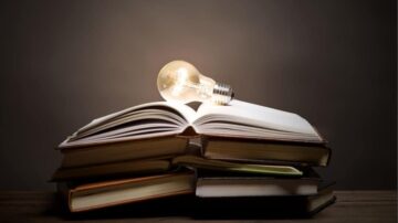 كتاب مفتوح مع مصباح كهربائي مضاء، موضوع على مجموعة من الكتب المغلقة. الخلفية داكنة، مما يبرز إضاءة المصباح. صورة تعبر عن الإبداع والمعرفة.