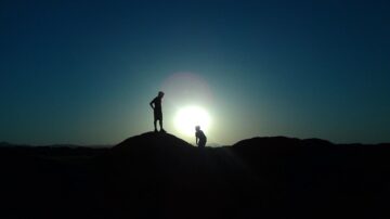 شخصان يتواجدان على قمة تلة مظلمة مع شمس الغروب خلفهما، مما يخلق تأثير ظل مثير. السماء زرقاء داكنة، تعكس جمال الطبيعة في لحظة هادئة.