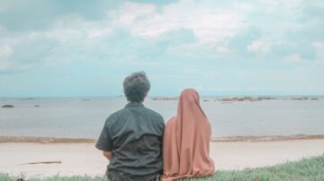 شخصان يجلسان على الشاطئ يراقبان البحر، الأول يرتدي قميصاً مزخرفاً والآخر يرتدي حجاباً. السماء ملبدة بالغيوم والمياه هادئة، مع ظهور صخور في الأفق.