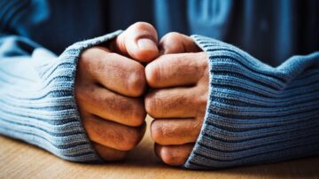 أيد مشدودة داخل أكمام سترة زرقاء، تعبير عن التوتر أو القلق، مع سطح خشبي في الخلفية.