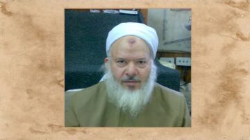 صورة لرجل مسن ذو لحية بيضاء، يرتدي عمامة بيضاء وعباءة، يجلس في مكان داخلي. تظهر خلفه سلالم ومكان مغطى بالسجاد. تعكس ملامحه الوقار والحكمة.