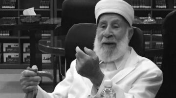 رجل مسن يرتدي جلباب أبيض وعمامة، يجلس في مكتبة ويُظهر تعبيرًا حكيمًا. يحمل عصا ويشير بإحدى يديه، محاطًا برفوف مليئة بالكتب.