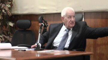 رجل مسن يرتدي بدلة رسمية يجلس على كرسي ويشير بيده إلى شيء خارج الصورة. خلفه يوجد مكتب خشبي وبعض النباتات.