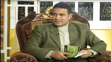 رجل يجلس في أريكة مريحة أثناء حديثه في برنامج تلفزيوني، يرتدي سترة رمادية وقميص بألوان فاتحة، holding a book in his hand. خلفية تحتوي على نوافذ مضاءة تعكس أجواء المدينة ليلاً.