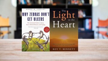 صورة لكتابين موضوعة على طاولة خشبية. الكتاب الأول هو "لماذا لا تصاب الزرافات بالقرحة" لمؤلفه روبرت م. سابولسكي، يتناول مواضيع التوتر والأمراض المرتبطة به. الكتاب الثاني هو "النور في القلب" للكاتب روي ت. بينيت، يقدم أفكاراً لتحسين الحياة اليومية. الخلفية تحتوي على أرفف كتب غير واضحة.