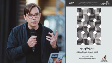 رجل يتحدث أمام جمهور، يحمل ميكروفونًا. خلفه جدارية ملونة وملفتة، بينما على الجانب الأيمن صورة لغلاف كتاب بعنوان "عصر نظام جديد". الكتاب يناقش التكنولوجيا والتصميم ومستقبل الإنسانية.