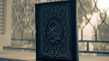 كتاب القرآن الكريم مفتوح مع تصميم مزخرف يظهر النص العربي. الخلفية ضبابية مع نافذة تضيء الضوء الطبيعي.